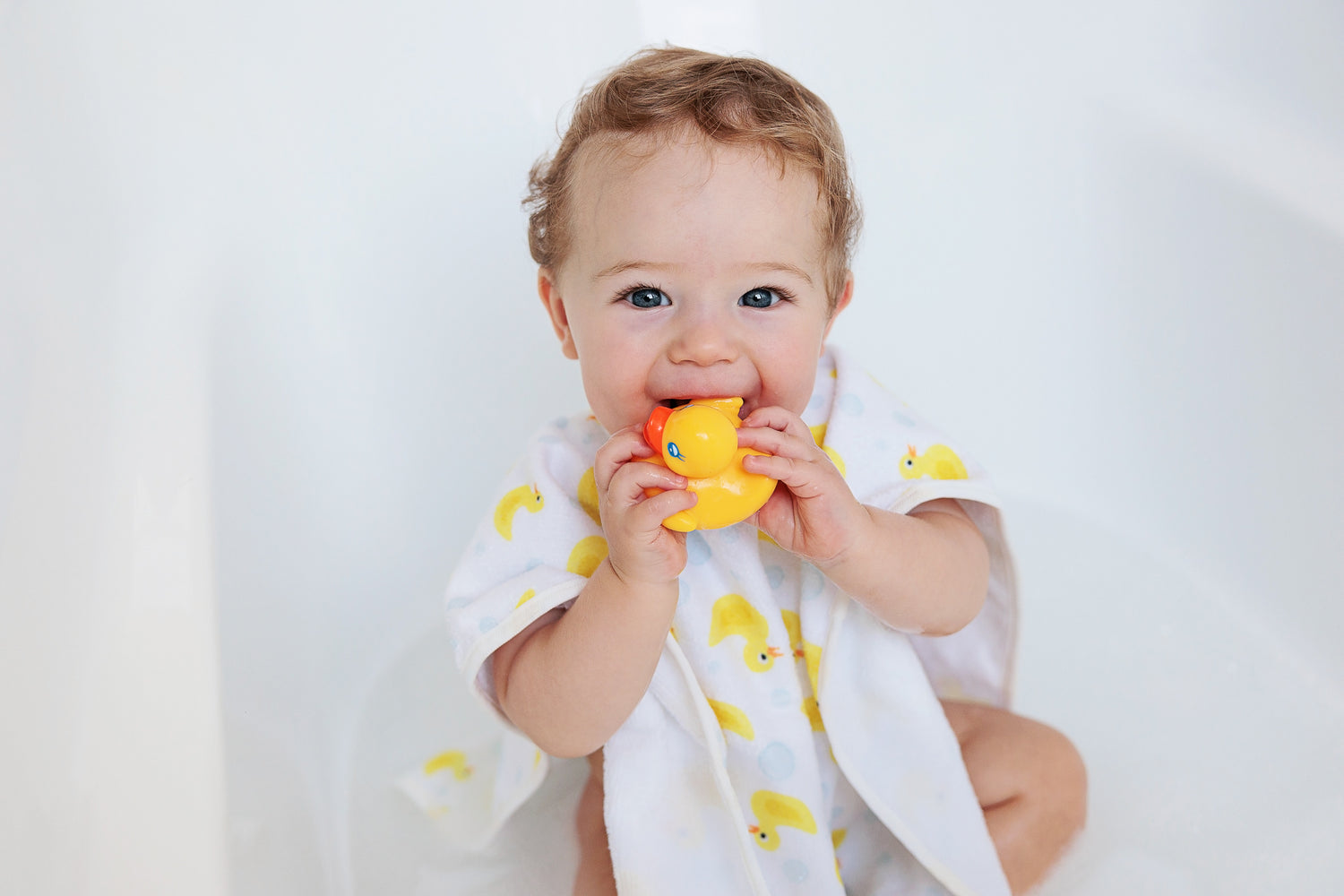 A little sandy blonde boy is being cheeky whilst he is chewing on a little yellow rubber bath duck. His blue eyes look directly into the camera. He sits in warm ankle deep bath water wearing a bath shirt covered in yellow ducks and little water bubbles. 