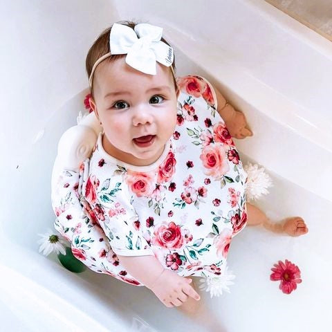 A little baby proudly looks up at her mum holding a white flower from her flower and milk bath. She is seated in a bath seat and wearing a bath shirt to keep her warm. She has a pretty white bow on her headband